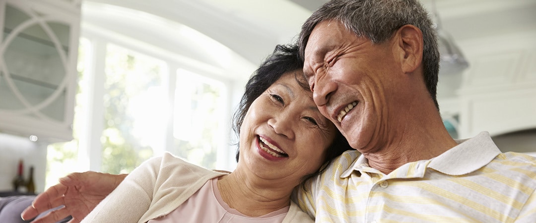 A senior man and woman sitting on a couch laughing together.