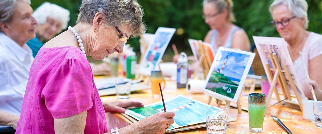 A senior woman outside painting on a canvas with other women. A senior woman outside painting on a canvas with other women.